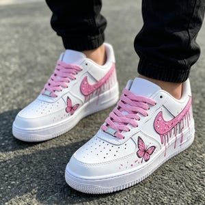 May include: White sneakers with pink accents, including the Nike swoosh, laces, and dripping paint effect. Features a butterfly design on the side. The shoes have a white sole and are worn by a person wearing black pants.