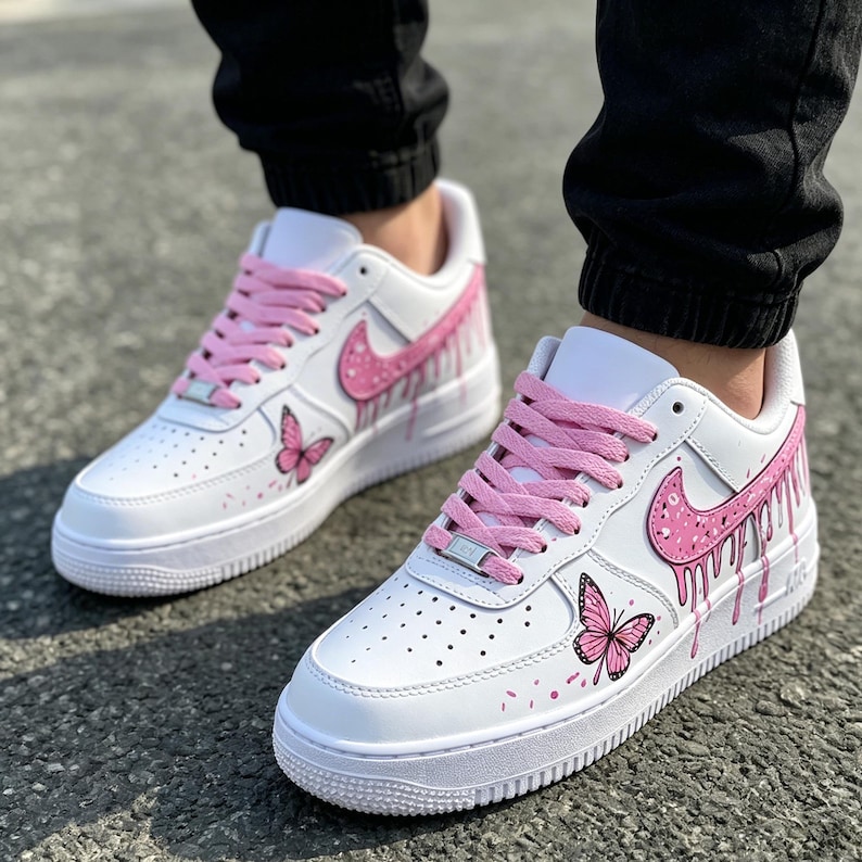 May include: White sneakers with pink accents, including the Nike swoosh, laces, and dripping paint effect. Features a butterfly design on the side. The shoes have a white sole and are worn by a person wearing black pants.
