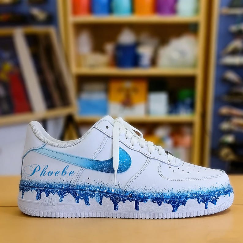 May include: White athletic shoe with a blue swoosh and custom blue paint details. The name "Phoebe" is written in cursive on the side. The shoe features a dripping paint design in shades of blue.