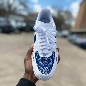 May include: White and blue athletic shoe with a paisley design on the toe and heel. The shoe has white laces and a blue Nike swoosh. The shoe is held in a person's hand.