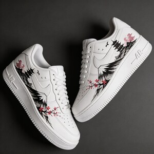 May include: White sneakers with a custom design featuring a Japanese landscape. The design includes a mountain, pagoda, and cherry blossoms in black, pink, and red. The shoes have white laces and soles, with "AR" printed on the side.