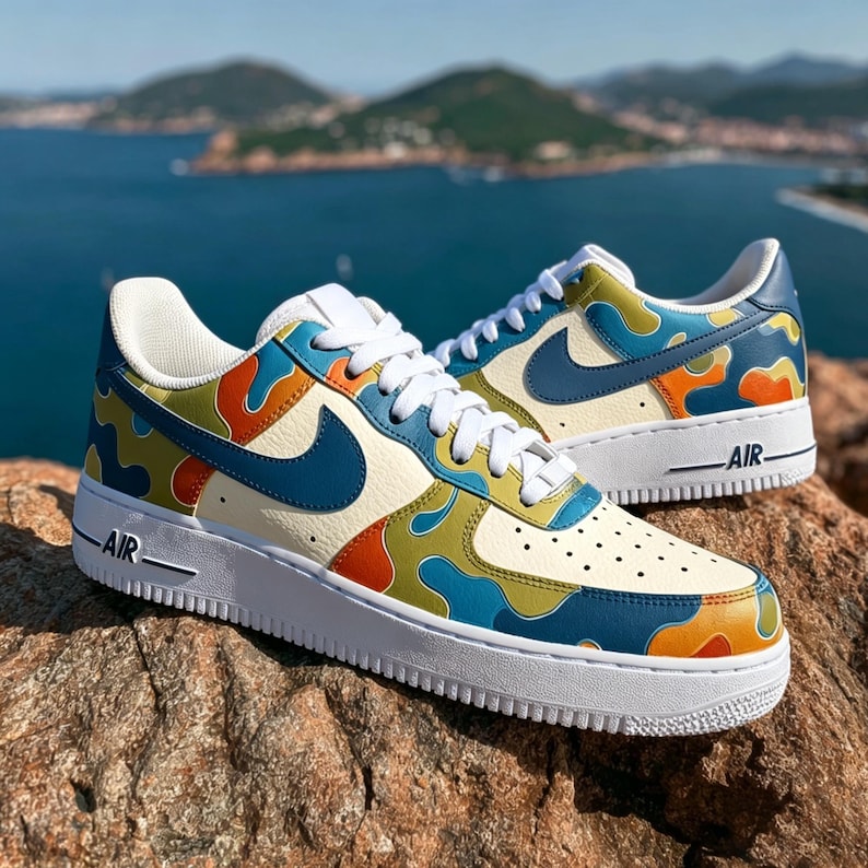 May include: A pair of custom Air Force 1 sneakers with a cream-colored base and a colorful camouflage pattern in blue, orange, and green. The shoes have white laces, a blue Nike swoosh, and a white sole. The word "AIR" is printed on the side.