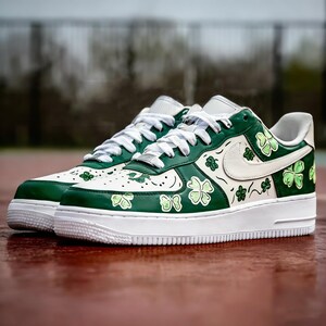 May include: Pair of white and green sneakers with shamrock designs. The shoes have a white sole, laces, and Nike swoosh. The green sections are decorated with shamrock patterns and the shoes are on a red surface.
