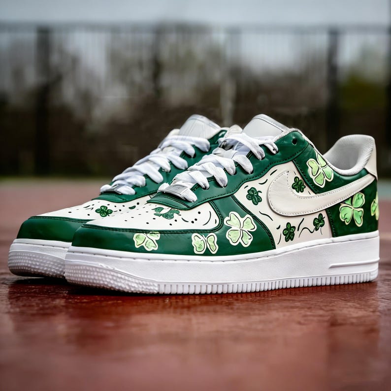 May include: Pair of white and green sneakers with shamrock designs. The shoes have a white sole, laces, and Nike swoosh. The green sections are decorated with shamrock patterns and the shoes are on a red surface.