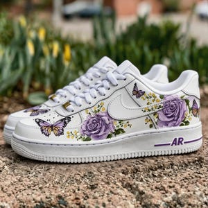 Op de afbeelding: Witte sneakers versierd met paarse rozen en vlindermotieven. De schoenen hebben een witte basis met een paarse zool en de letters "AR". Het ontwerp omvat paarse rozen, gele bloemen en vlinders.