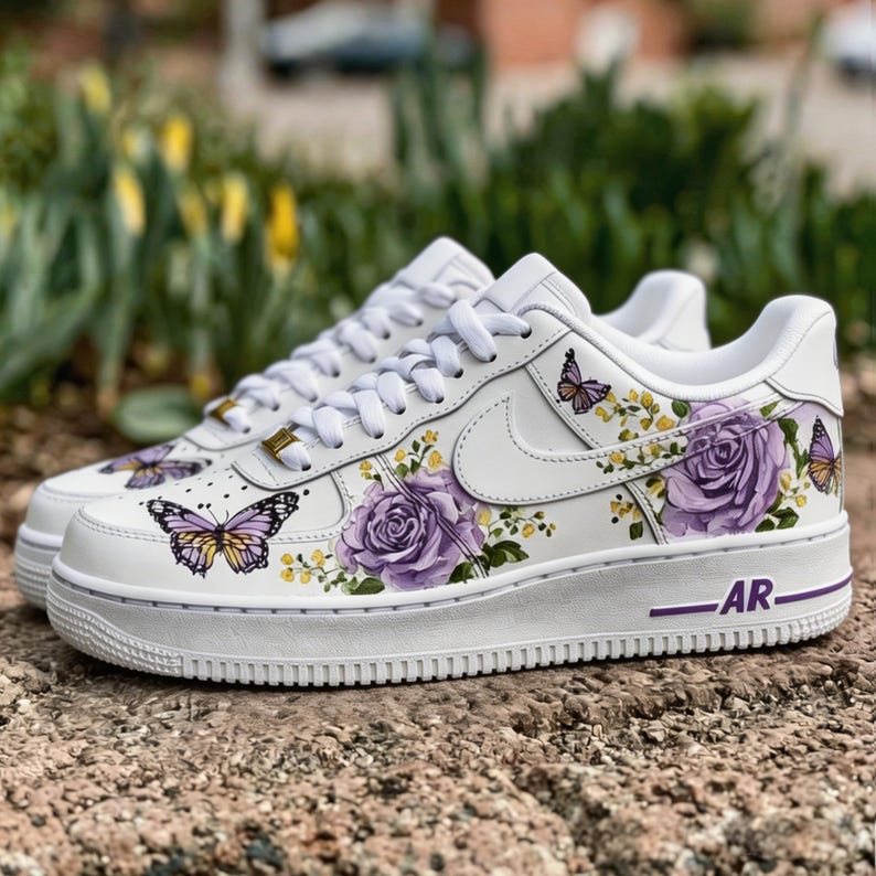 Op de afbeelding: Witte sneakers versierd met paarse rozen en vlindermotieven. De schoenen hebben een witte basis met een paarse zool en de letters "AR". Het ontwerp omvat paarse rozen, gele bloemen en vlinders.