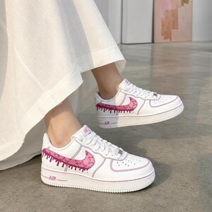 May include: White sneakers with pink accents. The shoes feature a pink swoosh design and dripping paint effect along the sides. The word "AIR" is printed on the midsole. The shoes are on a gray surface.