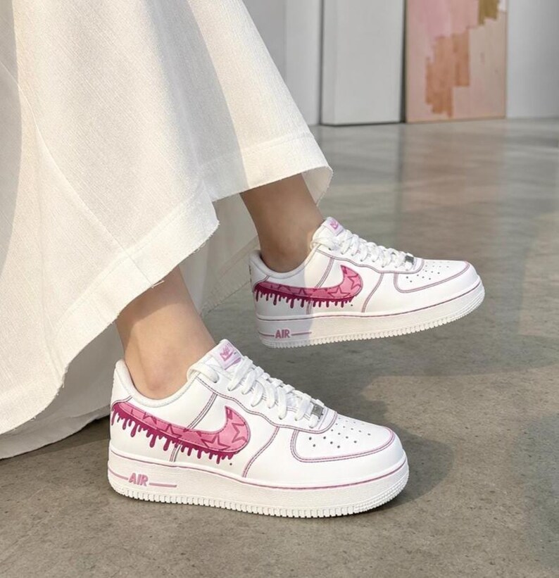 May include: White sneakers with pink accents. The shoes feature a pink swoosh design and dripping paint effect along the sides. The word "AIR" is printed on the midsole. The shoes are on a gray surface.