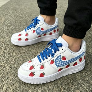 May include: White sneakers with blue laces and a strawberry design. The Nike swoosh is a blue and white gingham pattern. Red strawberries are scattered across the white leather upper. The soles are white.