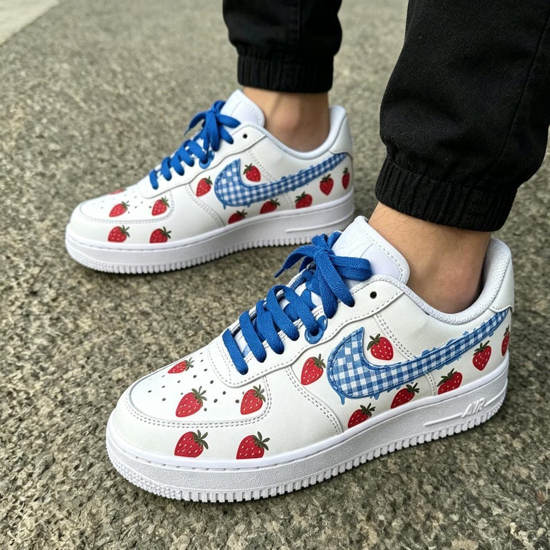 May include: White sneakers with blue laces and a strawberry design. The Nike swoosh is a blue and white gingham pattern. Red strawberries are scattered across the white leather upper. The soles are white.