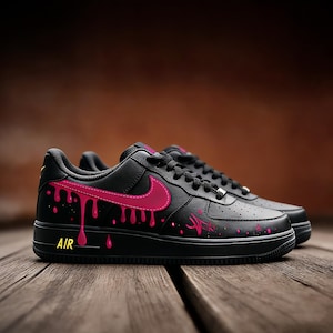 May include: Black Air Force 1 sneakers with pink dripping paint design. The Nike swoosh and accents are also pink. The word "AIR" is in yellow on the side of the sole. The shoes are on a wooden surface.