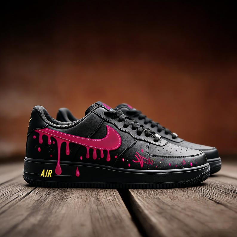 May include: Black Air Force 1 sneakers with pink dripping paint design. The Nike swoosh and accents are also pink. The word "AIR" is in yellow on the side of the sole. The shoes are on a wooden surface.