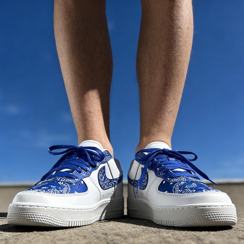 May include: White and blue sneakers with a paisley design on the toe box and heel. The sneakers have blue laces and a white sole. The Nike swoosh is blue and matches the laces and paisley pattern.
