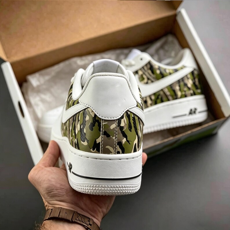 May include: White sneakers with a camouflage pattern on the side panels and heel. The shoes have a white sole and a black stripe. The sneakers are in a cardboard box.
