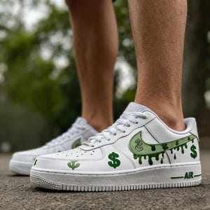 May include: White trainers featuring green dollar sign and dripping paint design. The Nike swoosh is green with a dollar sign graphic. The word "AIR" is printed in green on the midsole. The trainers are on a gray surface.