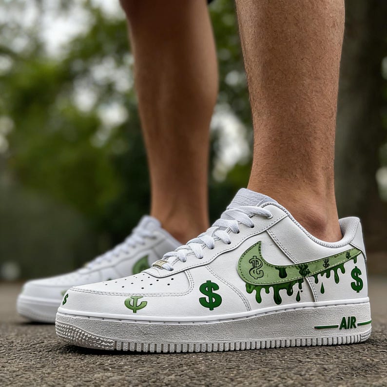 May include: White trainers featuring green dollar sign and dripping paint design. The Nike swoosh is green with a dollar sign graphic. The word "AIR" is printed in green on the midsole. The trainers are on a gray surface.