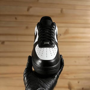May include: Black and white athletic shoe with a leather upper. The shoe features a white toe box with perforations, black laces, and a black sole. The iconic swoosh logo is visible on the side. The shoe is held by a gloved hand.