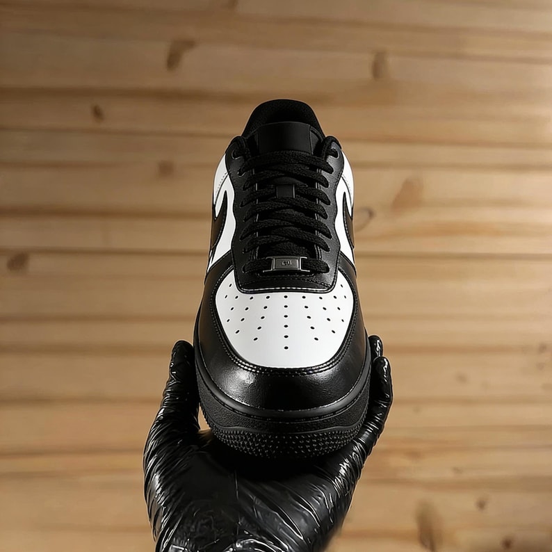 May include: Black and white athletic shoe with a leather upper. The shoe features a white toe box with perforations, black laces, and a black sole. The iconic swoosh logo is visible on the side. The shoe is held by a gloved hand.