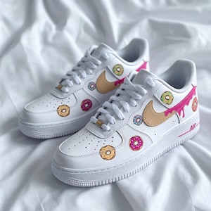 May include: White sneakers with a tan swoosh and donut graphics. The shoes feature pink, yellow, blue, and green donut designs. The laces are white, and the soles are white with a pink accent.