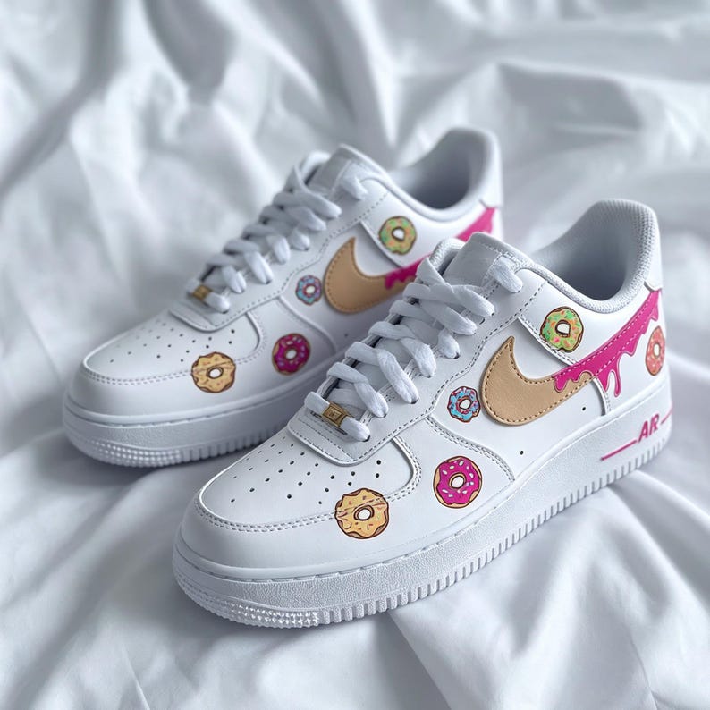 May include: White sneakers with a tan swoosh and donut graphics. The shoes feature pink, yellow, blue, and green donut designs. The laces are white, and the soles are white with a pink accent.