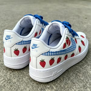 May include: White Nike Air Force 1 sneakers with red strawberry graphics and blue gingham accents. The shoes have blue laces and the Nike logo on the side and heel. The soles are white.