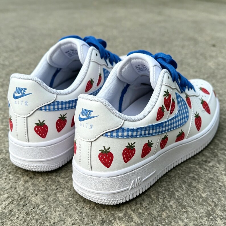 May include: White Nike Air Force 1 sneakers with red strawberry graphics and blue gingham accents. The shoes have blue laces and the Nike logo on the side and heel. The soles are white.