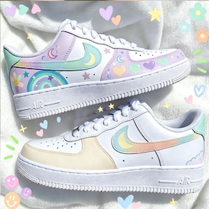 May include: A pair of white sneakers with pastel-colored accents. The shoes feature a design of moons, stars, hearts, and rainbows. The Nike swoosh is in a pastel color. The soles are white.