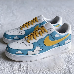 May include: A pair of custom-painted trainers. The shoes have a white base with light blue and white details, a yellow swoosh, and cloud graphics. The laces are yellow, and the soles are white.