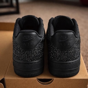 May include: A pair of black Nike Air sneakers viewed from the back. The shoes feature a textured, glittery black finish on the heel panels, with the Nike Air logo embossed in black on the leather heel tab. The soles are solid black rubber.
