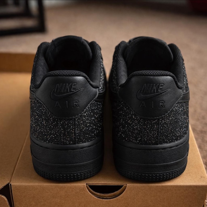 May include: A pair of black Nike Air sneakers viewed from the back. The shoes feature a textured, glittery black finish on the heel panels, with the Nike Air logo embossed in black on the leather heel tab. The soles are solid black rubber.