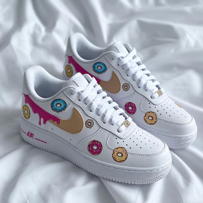 May include: White sneakers with custom donut designs. The shoes feature a tan Nike swoosh, pink dripping icing, and various donut graphics in pink, blue, and yellow. The word "AIR" is printed in pink on the side.