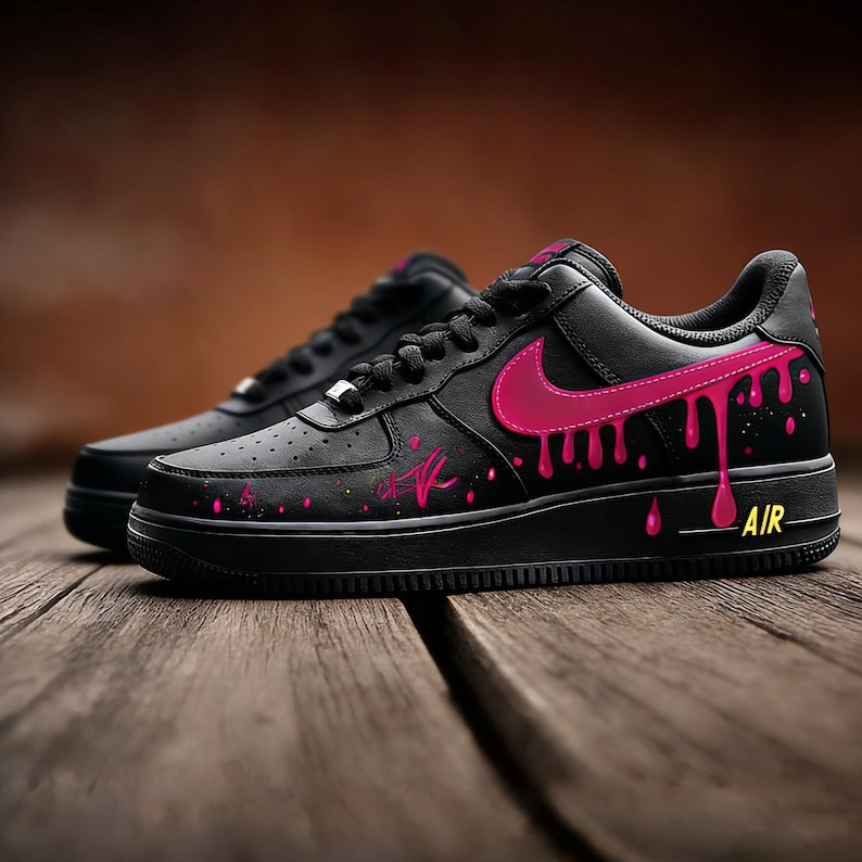 May include: Pair of black sneakers with a pink swoosh and dripping paint design. The shoes have a black sole and laces, with the word "AIR" in yellow on the side. The design includes pink splatters and a stylized signature.