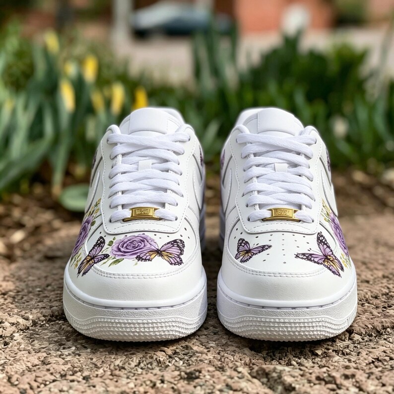 Op de afbeelding: Een paar witte sneakers met vlinder- en rozenmotieven. De schoenen hebben paarse en gele vlinders, een paarse roos en gouden details op de veters. De sneakers hebben witte veters en zolen.