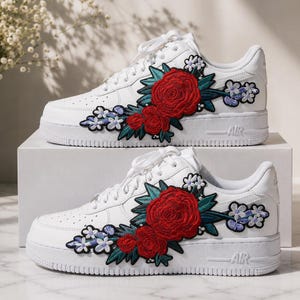 May include: A pair of white sneakers featuring embroidered floral patches on the side panels. The embroidery displays large red roses with dark green leaves and smaller light purple and white flowers, set against the smooth white leather upper.