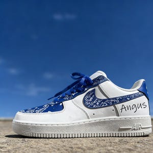 May include: White and blue Air Force 1 sneakers with a paisley pattern on the toe, swoosh, and heel. The laces and accents are blue. The name "Angus" is written in black on the side of the shoe. The sole is white.
