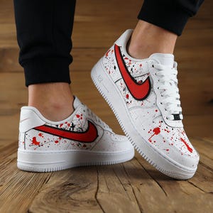 Może przedstawiać: Białe trampki z czerwonymi i czarnymi rozpryskami farby. Trampki mają czerwone logo Nike i białą podeszwę. Buty znajdują się na drewnianej powierzchni.