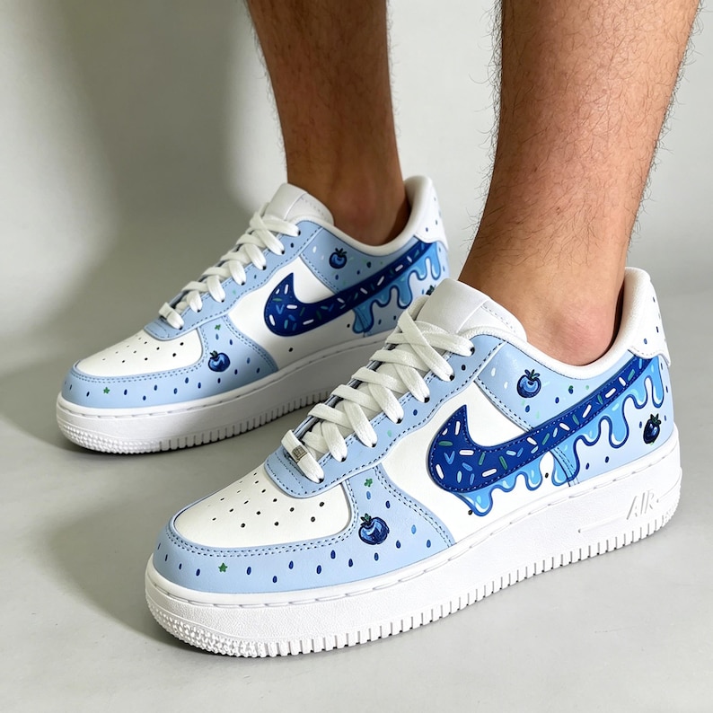 May include: White and light blue sneakers with a blueberry design. The shoes feature a white base with light blue accents, a dark blue swoosh, and a dripping blueberry design. The soles are white.