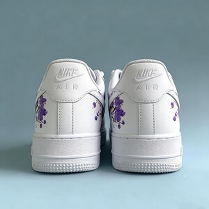 May include: Pair of white Nike Air Force 1 sneakers with purple floral accents on the heels. The Nike logo is visible on the back of each shoe. The sneakers have a classic design with a white sole.