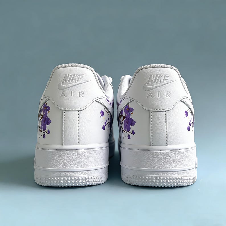May include: Pair of white Nike Air Force 1 sneakers with purple floral accents on the heels. The Nike logo is visible on the back of each shoe. The sneakers have a classic design with a white sole.