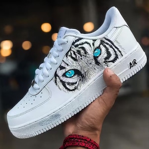 May include: White leather sneaker with a tiger eye design. The sneaker features a detailed illustration of a tiger's face with striking blue eyes. The word "AIR" is printed on the side of the shoe. The laces are white.