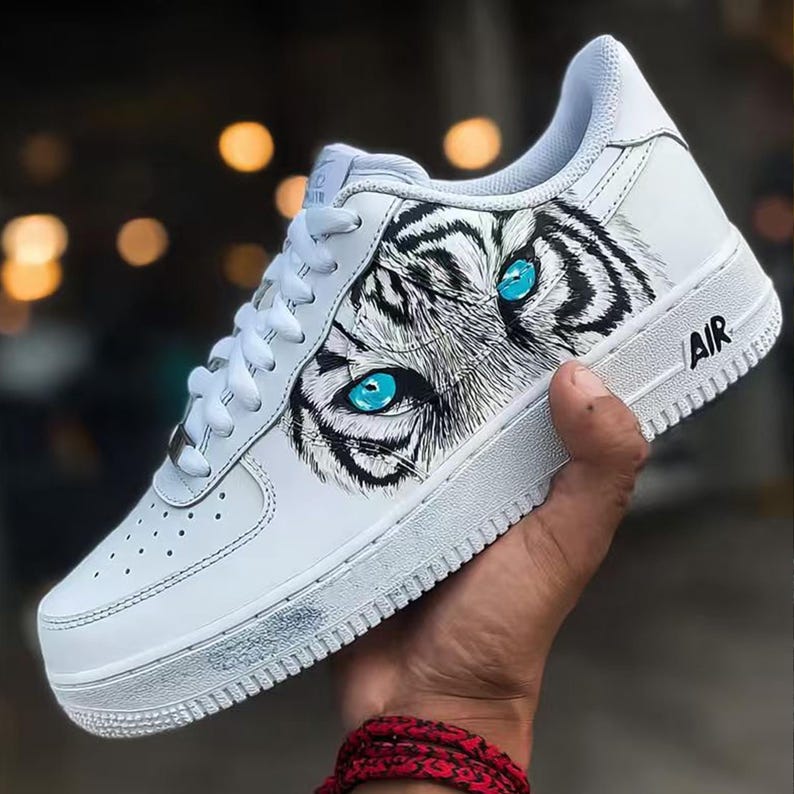 May include: White leather sneaker with a tiger eye design. The sneaker features a detailed illustration of a tiger's face with striking blue eyes. The word "AIR" is printed on the side of the shoe. The laces are white.