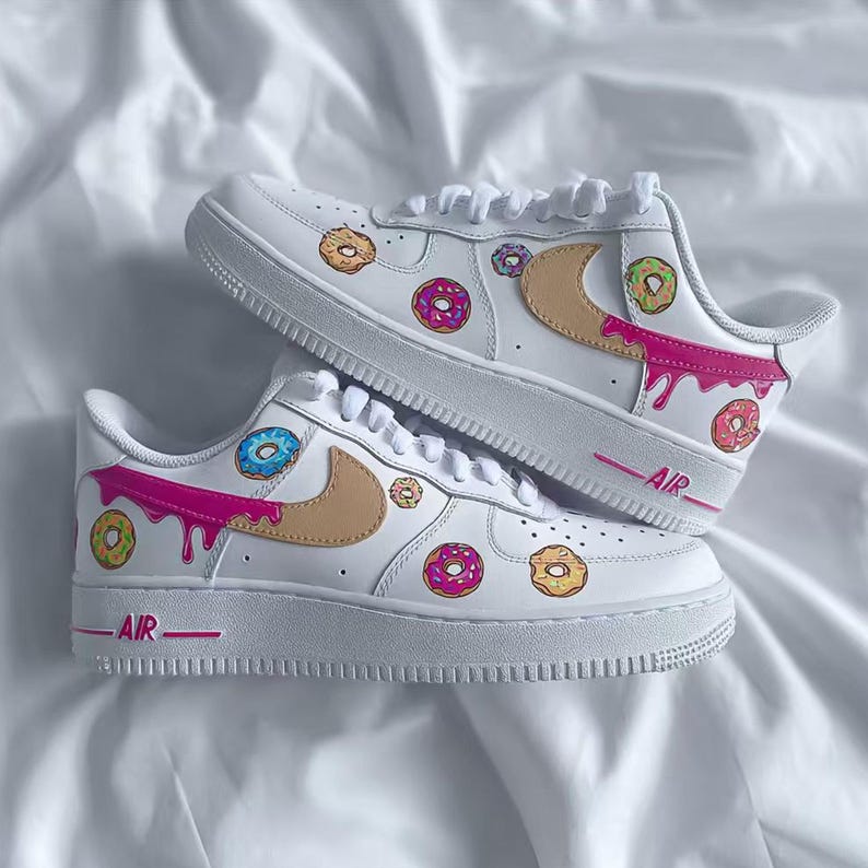 May include: White sneakers with a playful donut design. The shoes feature a tan swoosh, pink dripping accents, and colorful donut graphics. The word "AIR" is printed in pink on the side of the sole.