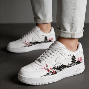 May include: White sneakers with a Japanese-inspired design. The shoes feature a black and gray mountain range, a pagoda, and cherry blossoms in pink and red. The design is on the side of the shoe.