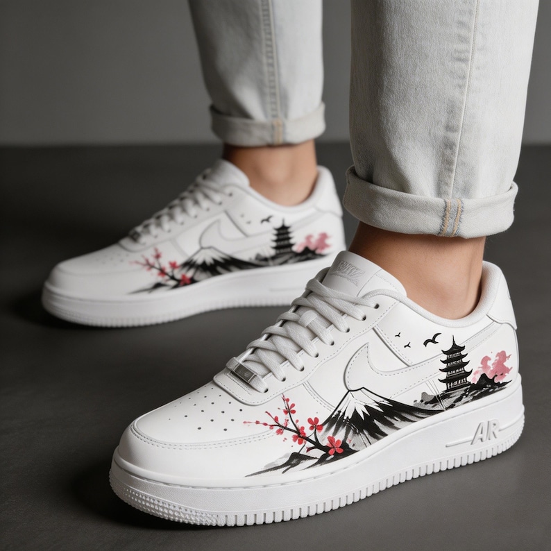 May include: White sneakers with a Japanese-inspired design. The shoes feature a black and gray mountain range, a pagoda, and cherry blossoms in pink and red. The design is on the side of the shoe.