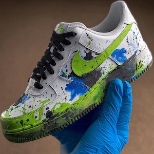 May include: White and black Air Force 1 sneaker with green and blue paint splatter design. Features a green Nike swoosh, black laces, and the word "AIR" on the side. The shoe is held by a person wearing a blue glove.