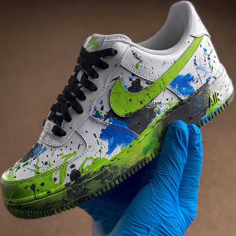 May include: White and black Air Force 1 sneaker with green and blue paint splatter design. Features a green Nike swoosh, black laces, and the word "AIR" on the side. The shoe is held by a person wearing a blue glove.