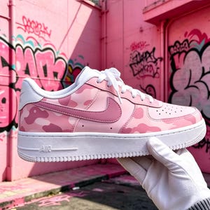May include: Pink camouflage Nike Air Force 1 sneakers with a white sole and laces. The shoe features a pink camouflage pattern on the upper, a pink Nike swoosh, and the word "AIR" on the sole.