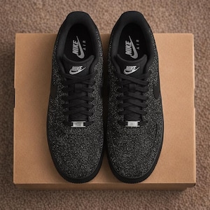 May include: A pair of black Nike Air Force 1 sneakers featuring a glittery, textured upper, black laces, and a silver metallic AF1 lace dubrae. The interior lining is black with white Nike Air branding on the insole and tongue label.