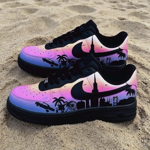 May include: Pair of black and multi-colored sneakers with a city skyline design. The shoes feature a gradient of pink, purple, and blue, with black accents and a black sole. The design includes palm trees, a Ferris wheel, and a car.