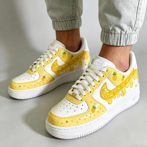 May include: White and yellow sneakers with a pineapple design. The shoes feature a white base with yellow accents, including a dripping pineapple design on the Nike swoosh and sides. Small pineapple and star details are scattered across the shoe.